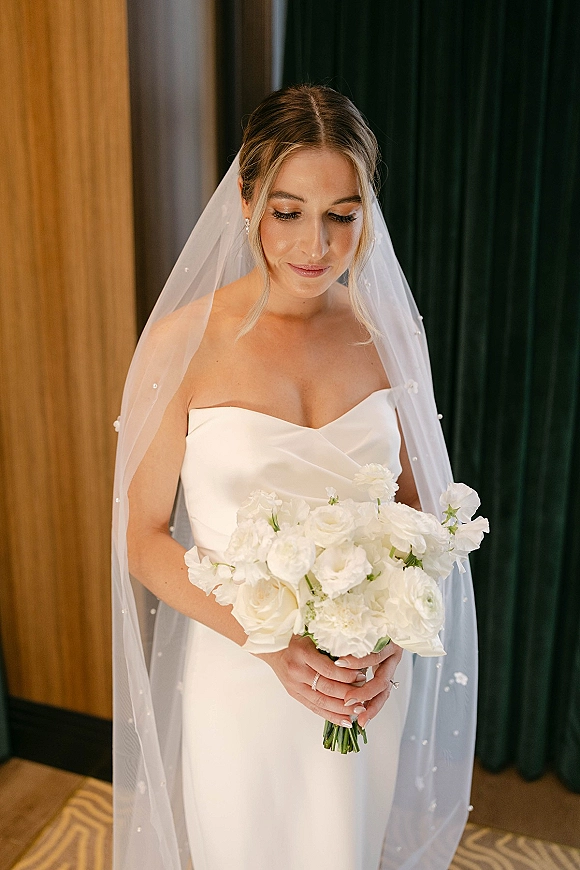 Bridal portrait of a bride holding bouquet in a strapless wedding dress, looking down with a pearl veil before indoor curtains and wood paneling