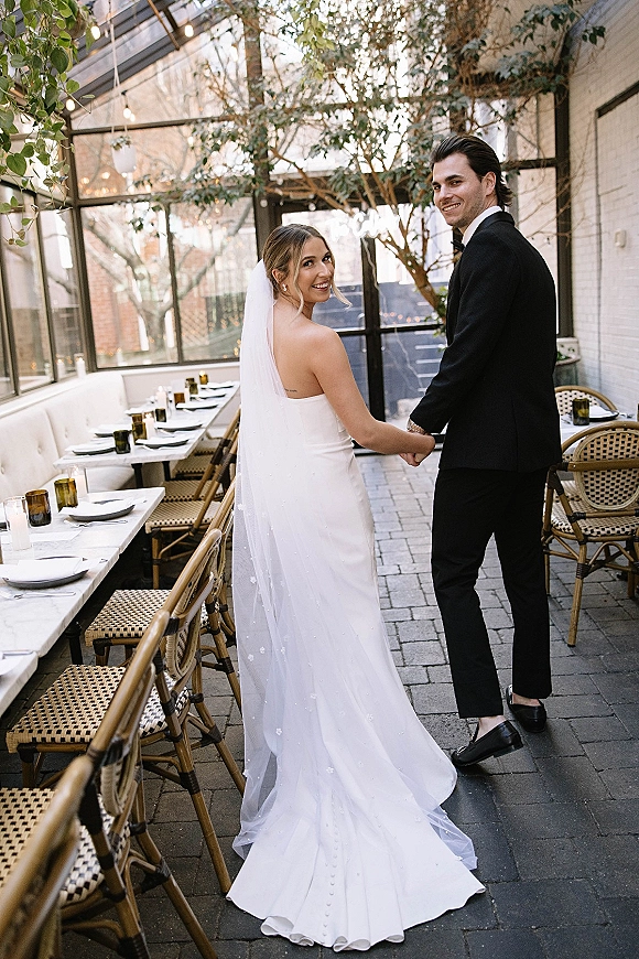Couple portrait of bride and groom holding hands, looking back in a glass conservatory with veil train, tuxedo, and string lights overhead