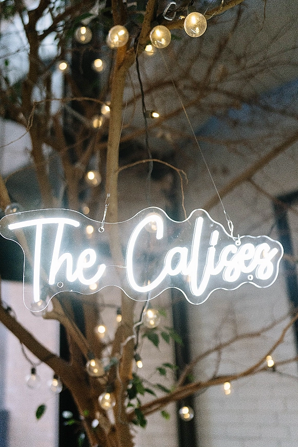 Wedding neon sign glowing in white script on a clear acrylic backing with string lights, hanging among tree branches by a building window at night
