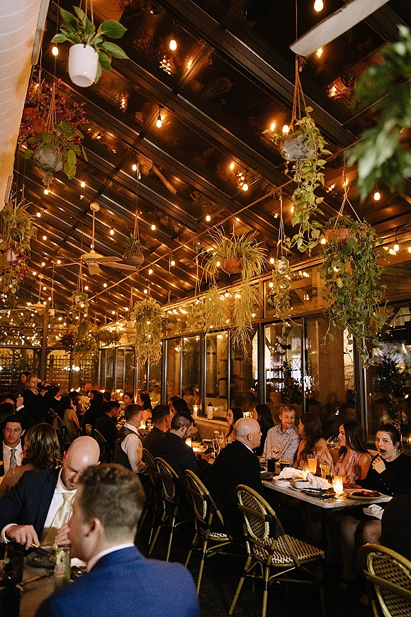 Reception dinner with long tables set with glassware and candle votives under string lights and hanging plants in a glass-ceiling greenhouse at night