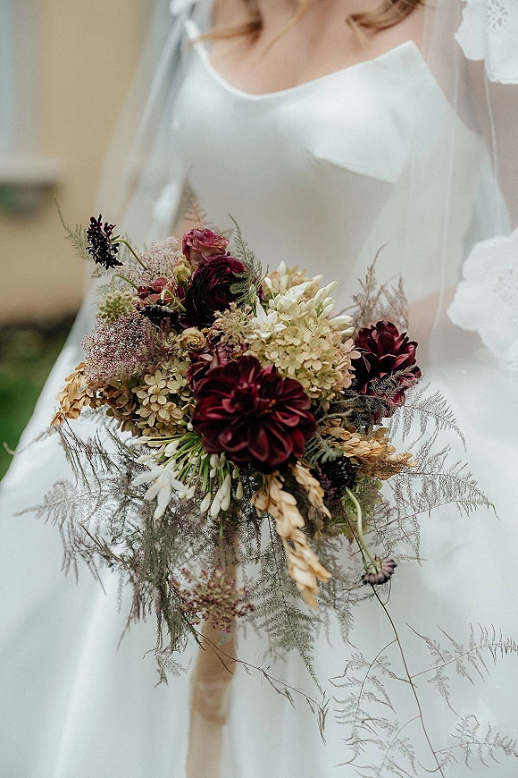 Bridal bouquet with burgundy wedding bouquet tones, cream hydrangea, dried florals, and greenery held by bride in veil on outdoor walkway