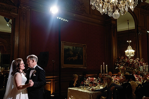 First dance as bride in veil and groom in tuxedo sway beneath a chandelier, surrounded by candlelit banquet tables and guests