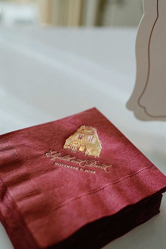 Wedding napkins with embossed gold foil monogram, stacked on a white tabletop beside a minimalist place card silhouette