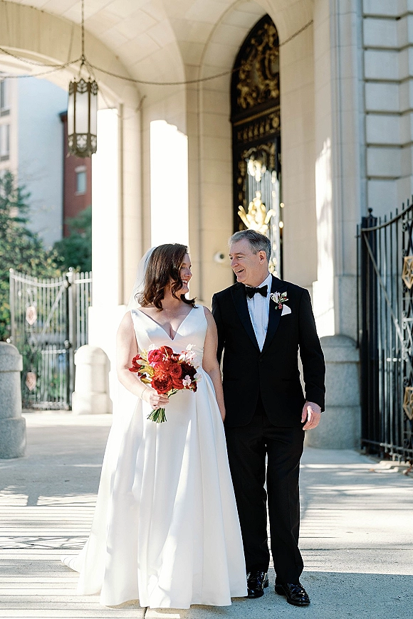 Couple portrait of bride and groom walking, her red bouquet and veil flowing as they smile outside a stone building with arched doorway
