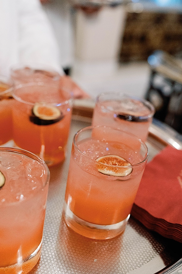 Wedding signature cocktail with pink drinks in glasses, fig garnish and ice on a tray, set at an indoor wedding cocktail bar with blurred decor