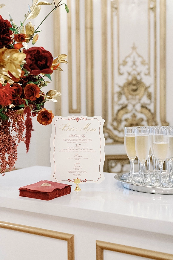 Wedding bar menu and signature drinks sign on a stand with champagne flutes, burgundy napkins, and florals at a white-and-gold bar counter