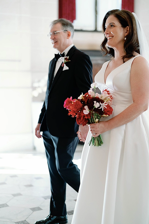 Bride walking down aisle holding a red and blush bouquet beside her father in a tuxedo, veil flowing in a bright hallway with windows