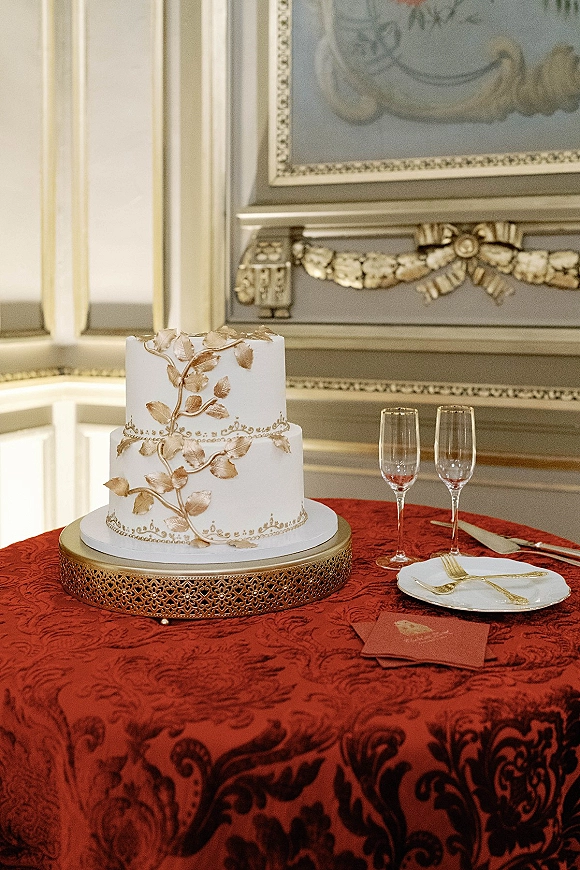 Wedding cake two tier wedding cake with gold leaf icing on a stand atop red damask tablecloth, champagne flutes and knife set nearby