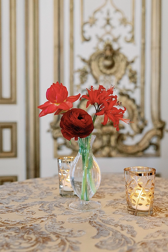 Reception tablescape with a wedding table centerpiece of red flowers in clear bud vases, votive candles in gold holders on damask cloth.
