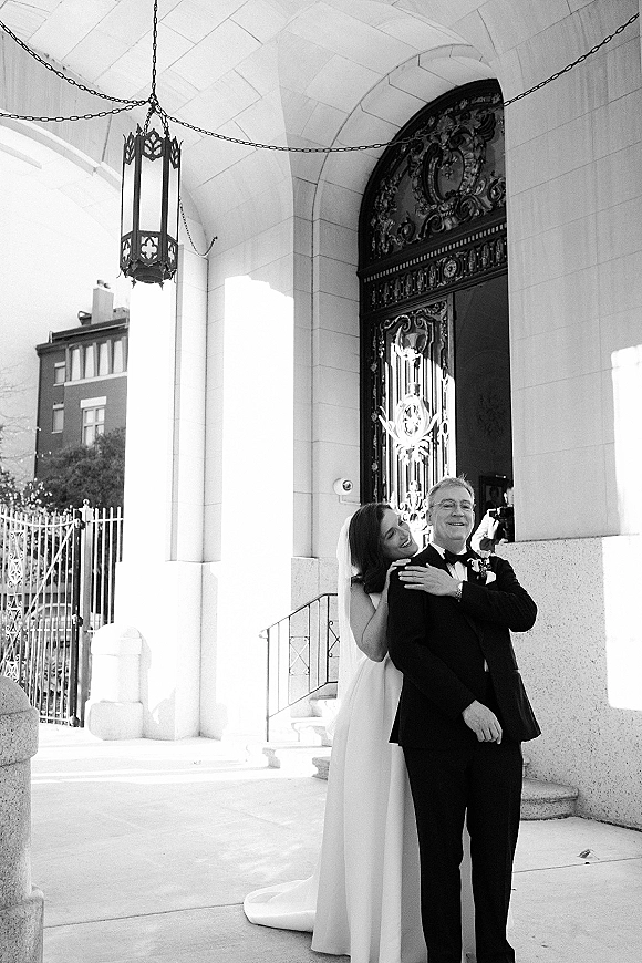 Couple portrait in black and white, bride hugging groom from behind with veil and tuxedo, posed on steps by a stone archway doorway