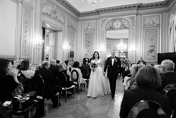 Wedding recessional as bride and groom walk hand in hand down the aisle, bride holding bouquet in an ornate ballroom with chandelier and guests seated