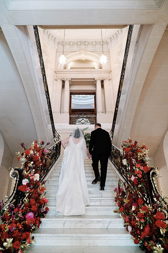 Couple portrait of bride and groom walking upstairs on a grand marble staircase, veil and tux, under chandeliers with burgundy florals