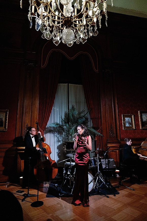 Wedding band playing jazz with a female vocalist in a red satin gown on a chandelier-lit stage in a wood-paneled ballroom