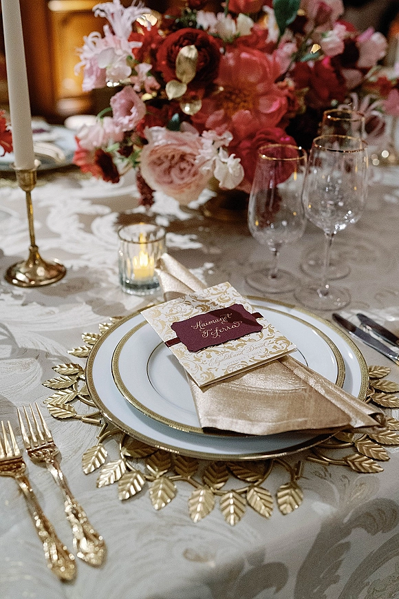 Wedding tablescape with reception place setting featuring gold charger plates, lush floral centerpiece, candles, and calligraphy menu on patterned linen