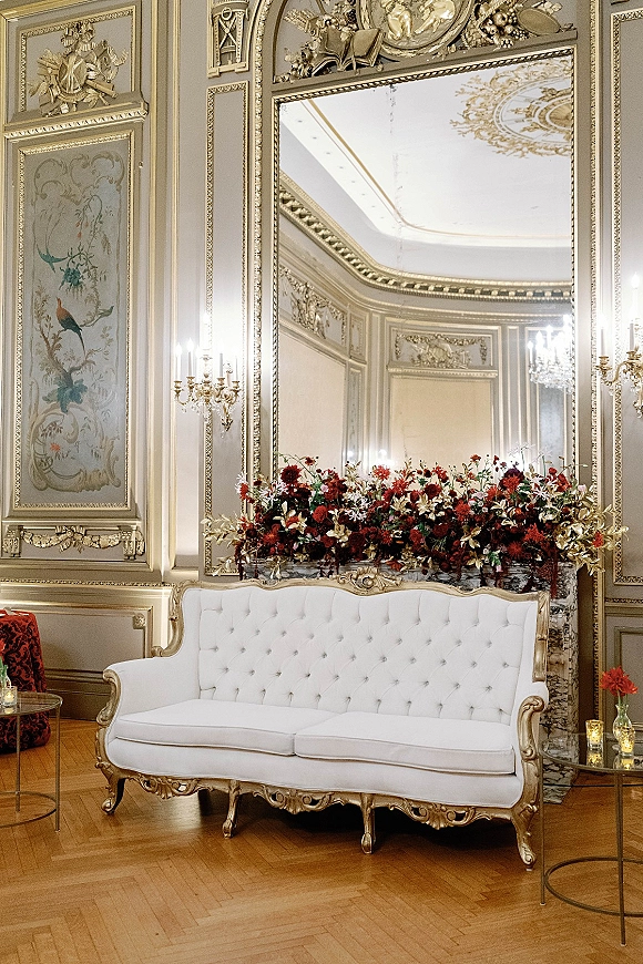 Wedding lounge seating with a white tufted sofa, gold ornate frame, red florals, and candlelit votives before a mirrored ballroom backdrop