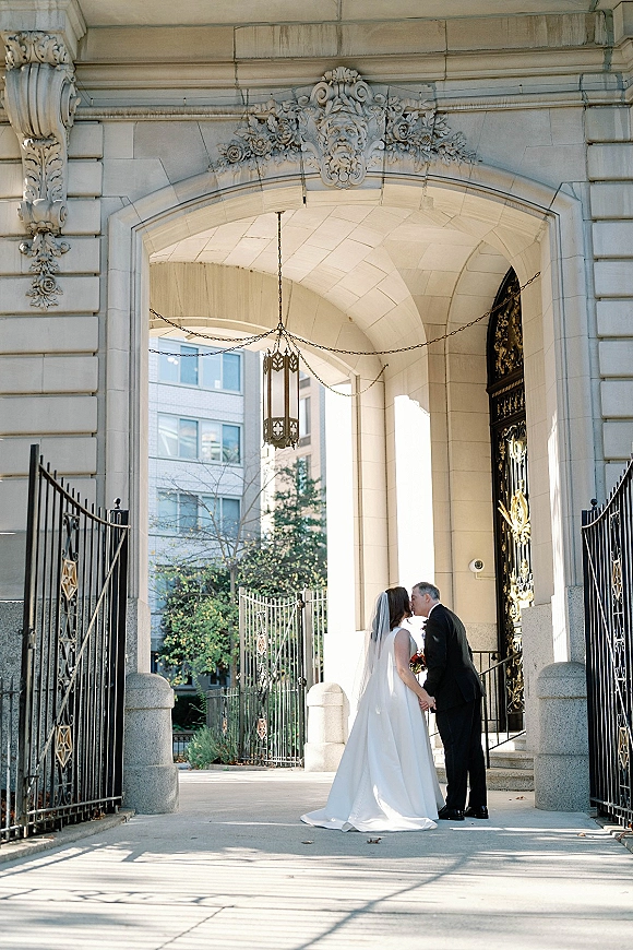 Wedding kiss portrait of bride and groom kissing under a stone archway, her long veil and bouquet framed by iron gates and lantern