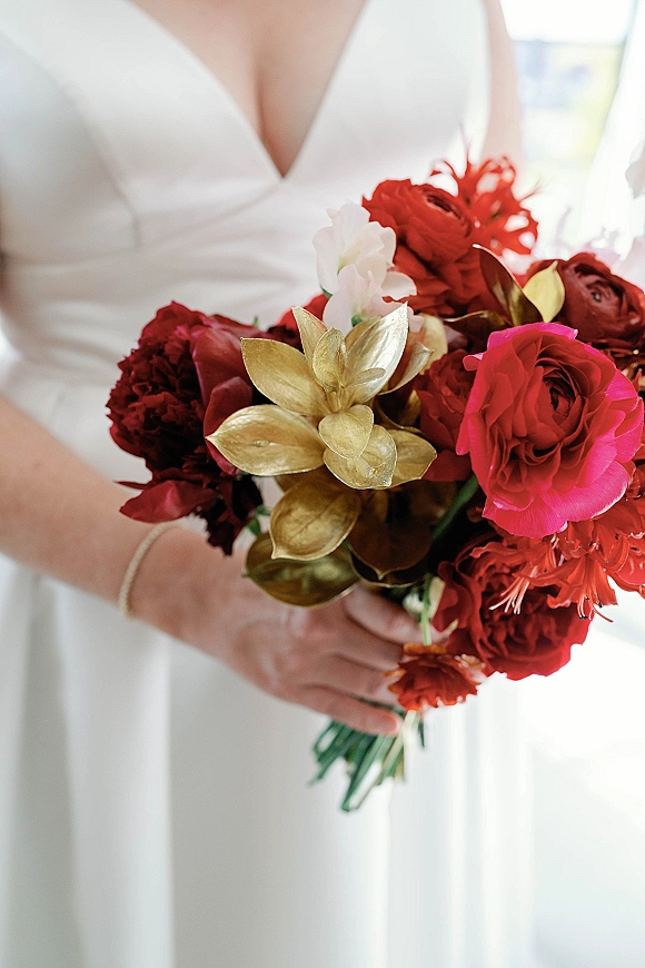 Bridal bouquet with red wedding bouquet blooms, pink rose and gold leaves held against a white gown in bright indoor window light