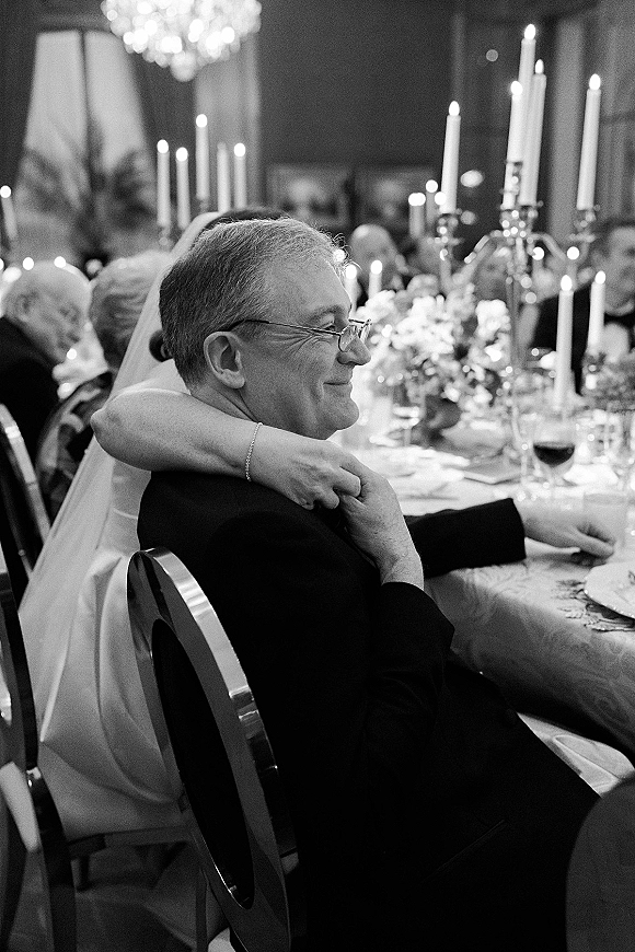 Wedding reception moment of the bride hugging groom seated at the head table under chandelier light, with taper candles and guests behind them