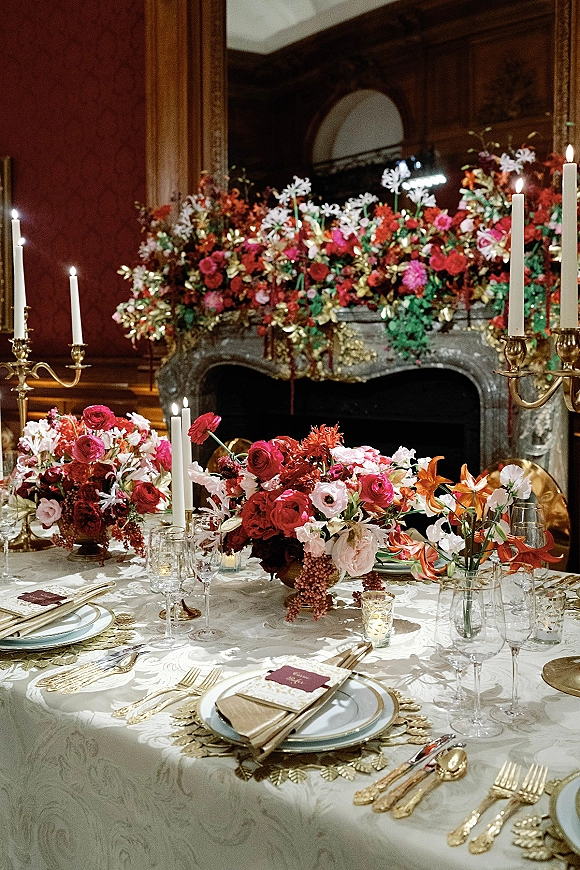 Reception tablescape with a wedding table centerpiece of red and pink florals, brass candelabras and taper candles before an ornate fireplace.