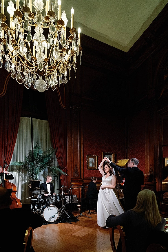 First dance as the bride twirls in her wedding dress with the groom in a suit beneath a chandelier, guests seated in a wood-paneled ballroom