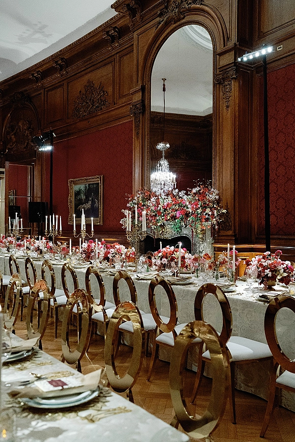 Reception tablescape with a long banquet table wedding setup, gold candelabras and taper candles, lush florals, and a crystal chandelier overhead