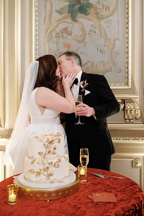 Wedding kiss as bride in veil and groom in tuxedo lean in by a candlelit wedding cake table with champagne flute in ornate reception room