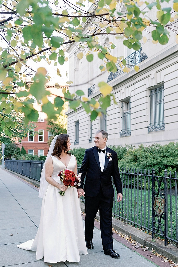 Couple portrait of bride and groom walking hand in hand, bride with burgundy bouquet and veil beside wrought iron fence and hedges