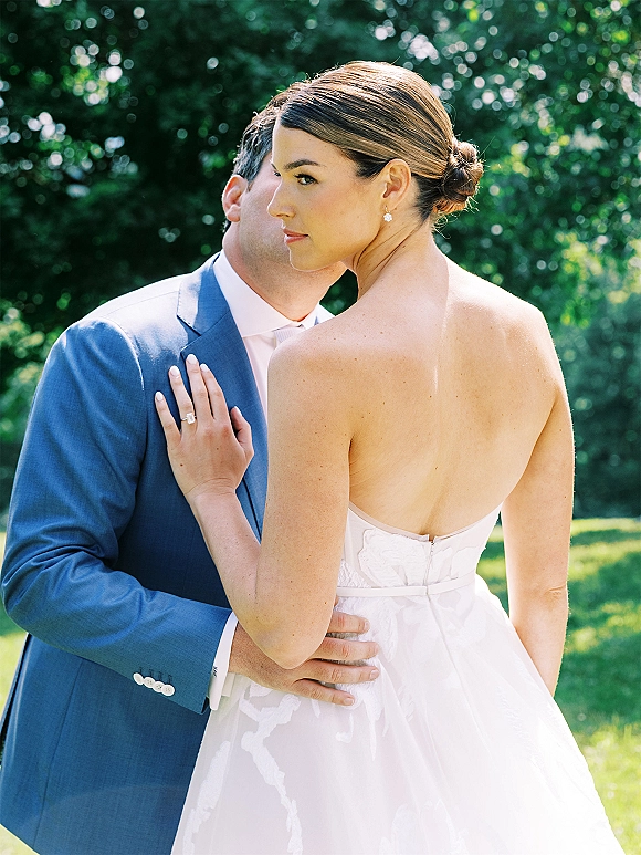 Couple portrait of bride looking over shoulder as groom embraces her, showing engagement ring and bands, in sunlit green trees backdrop