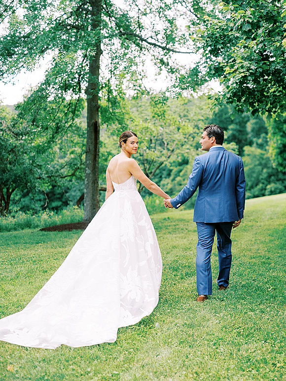 Couple portrait of bride and groom walking away holding hands on a grassy lawn with hills and trees, her dress train flowing