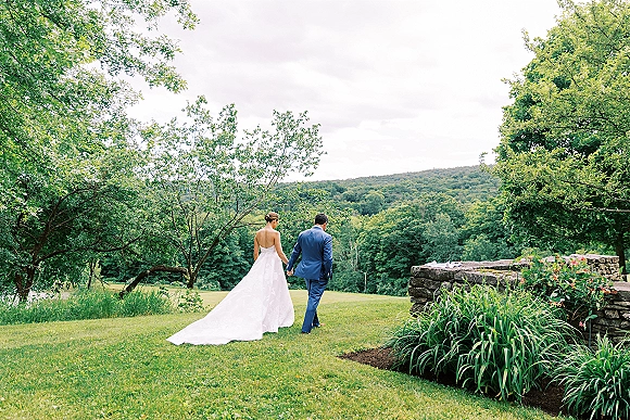 Couple portrait of bride and groom walking away hand in hand on grass, her strapless dress with long train, his blue suit near stone wall