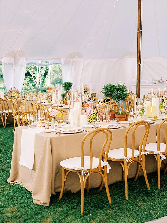 Reception tablescape with long banquet table setup featuring neutral linens, pastel florals and pillar candles under a white tent with string lights