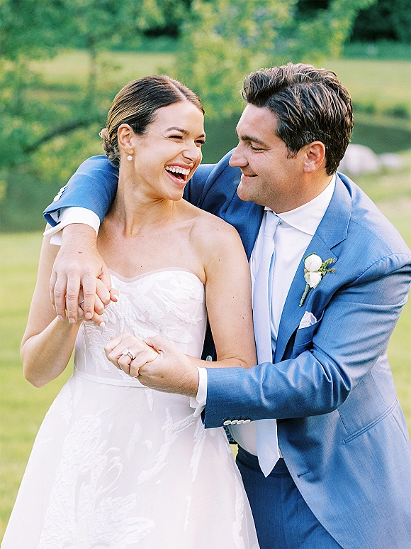 Couple portrait of a laughing bride and groom embracing, her strapless dress and his blue suit, on a green lawn by trees and pond