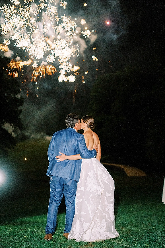 Wedding kiss portrait of bride in a strapless gown and groom in a blue suit under fireworks on a lawn at night with trees