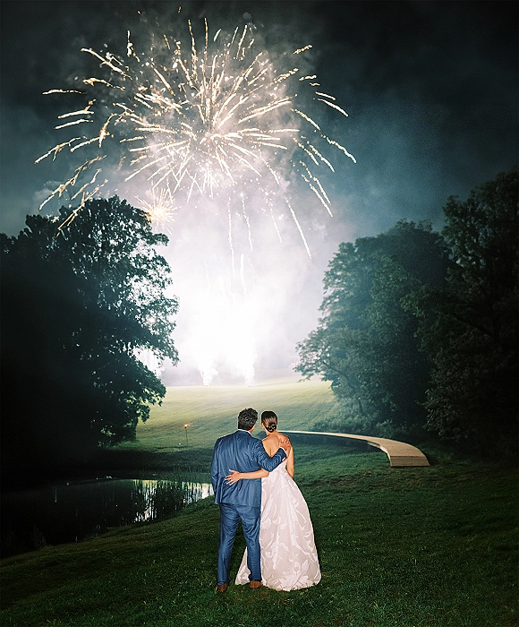 Wedding fireworks photo of bride in strapless gown and groom in blue suit watching bursts over a pond and footbridge at night