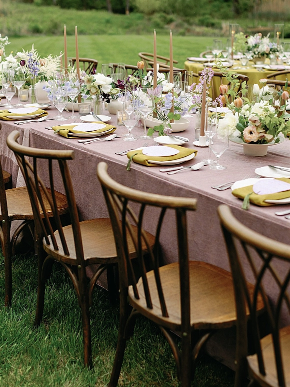 Reception tablescape with mauve tablecloth, olive green napkins, taper candles, bud vase florals, and glassware on a garden lawn