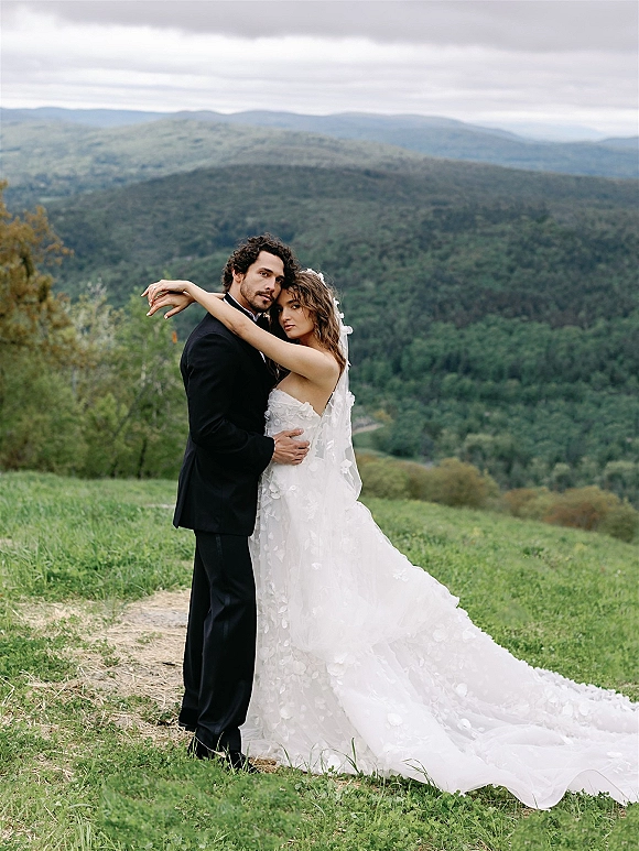 Couple portrait of bride and groom embrace, her lace applique gown and veil flowing in a grassy field with forested mountains under clouds