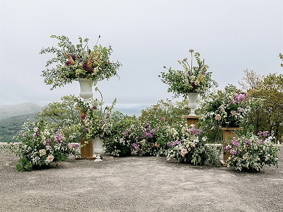 Wedding ceremony flowers with lush greenery garlands and pedestal urns on wooden plinths at a mountain overlook under cloudy skies