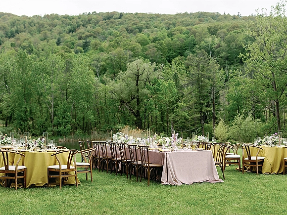 Outdoor reception tablescape with round and long banquet tables, pastel florals and taper candles on a lakeside lawn with forest hills backdrop