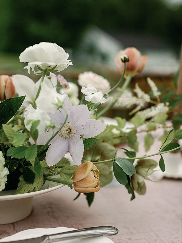 Wedding centerpiece with a low floral centerpiece of white flowers and peach tulips in a bowl vase on a blush linen table set outdoors