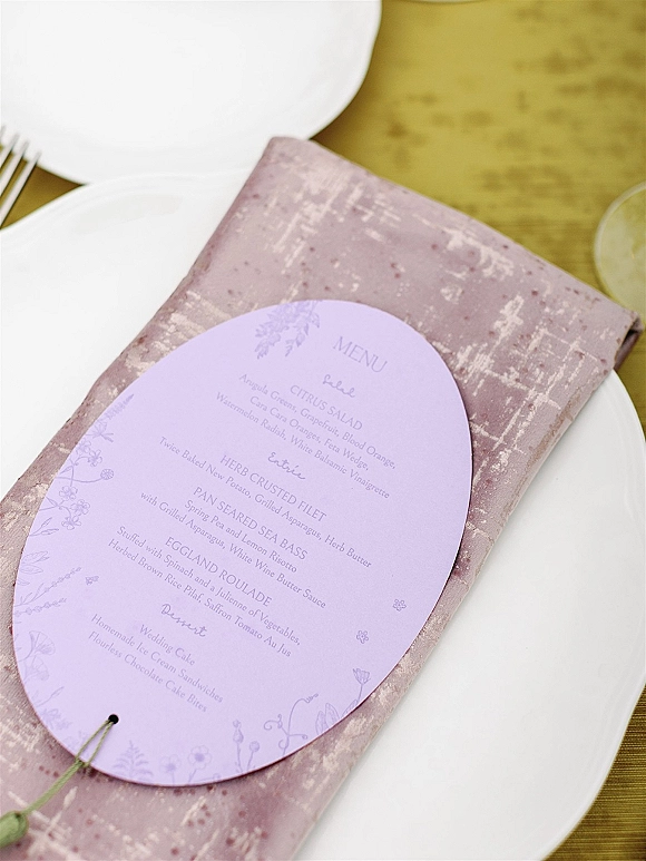 Wedding menu card in lavender, oval and tied with green string, rests on a mauve napkin by white plates and fork on gold tablecloth