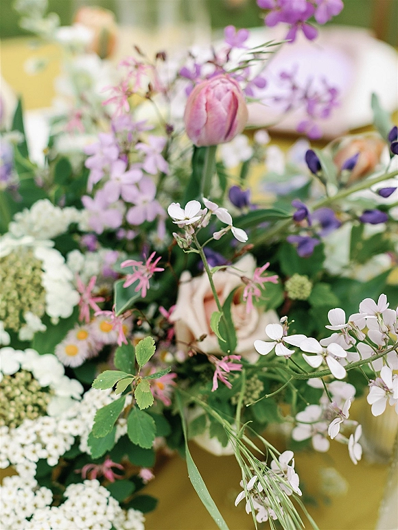 Wedding centerpiece with pastel blooms, featuring a pink tulip and peach rose amid purple flowers, white blossoms, and greenery on a softly blurred table