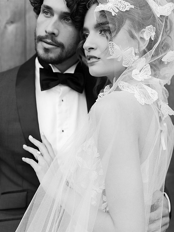 Couple portrait in a black and white wedding portrait style, bride and groom embrace with lace veil and bow tie by a wood wall