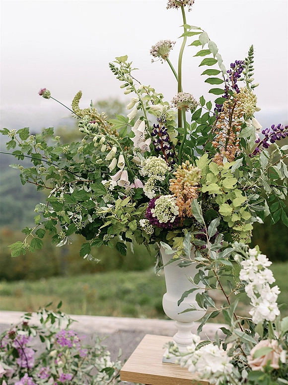 Wedding floral arrangement in a white urn vase with wildflowers, greenery, foxglove and purple blooms on a wooden pedestal outdoors