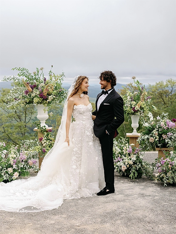 Couple portrait of bride in a strapless floral appliqué gown and long veil with groom in tuxedo under floral arch, mountains behind