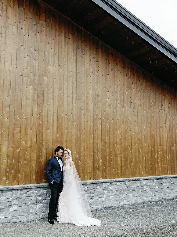 Couple portrait of bride in a lace wedding dress leaning on groom in tuxedo beside a rustic wood-and-stone wall under an overhang
