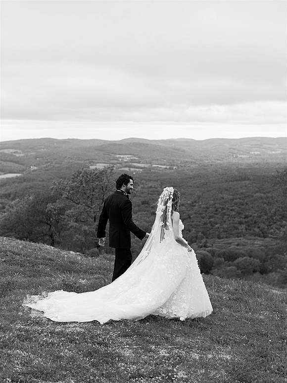 Couple portrait of bride and groom holding hands, walking away on a grassy hill with mountain view, her long train and veil flowing