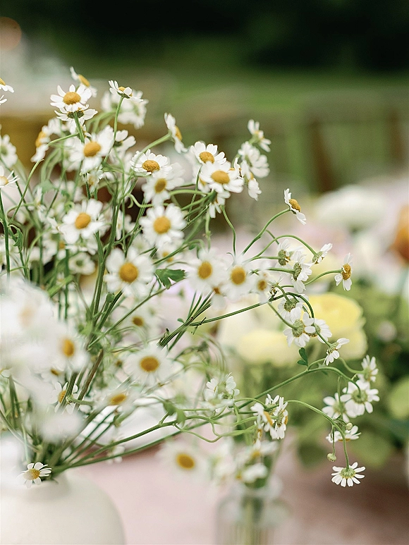 Wedding floral centerpiece with daisies and greenery in a glass vase on a white tablecloth, set on an outdoor garden reception table
