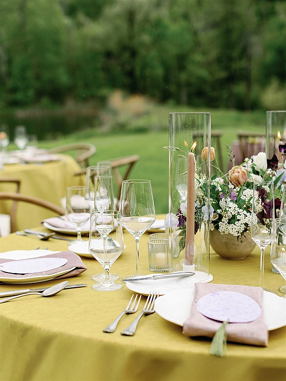Reception tablescape with a mustard tablecloth, blush napkins, menus, and taper candles in glass cylinders on an outdoor garden lawn