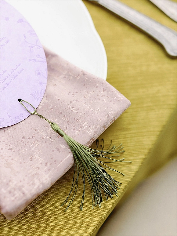 Wedding invitation suite flat lay featuring a round invitation card with blush envelope and green tassel on a white plate over gold linen