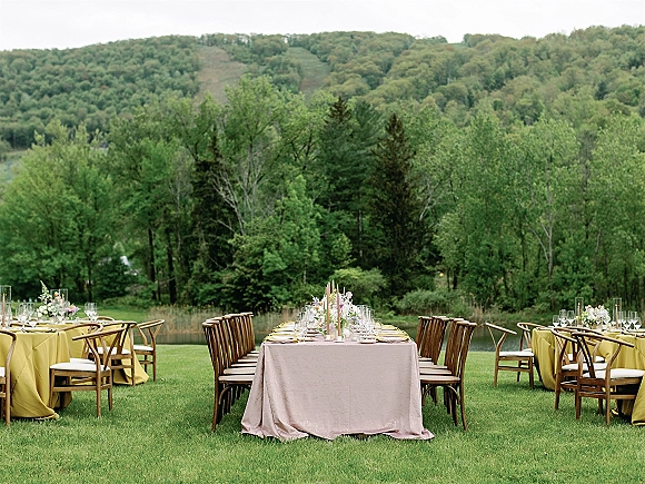 Outdoor reception tablescape with a long banquet table, floral centerpieces, and taper candles in glass hurricanes on a lakeside lawn